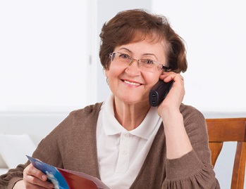 senior woman wearing a white polo and brown sweater sitting in a wooden chair smiling while talking on the telephone