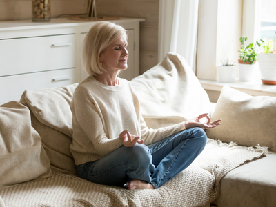older woman wearing cream sweater, meditating on couch
