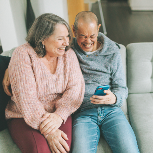 Older-adult couple sitting on sofa at home, laughing while looking at cellphone