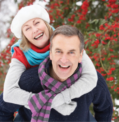 older-adult couple dressed in warm winter clothing, smiling for photo outside, background of winter trees and snow